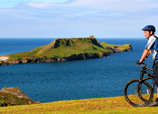 Worms Head South Wales UK. View from the cliffs across the water to Worms Head geological feature. Two unknown cyclists on mountain bikes wearing helmets taking in the view. Blue sky